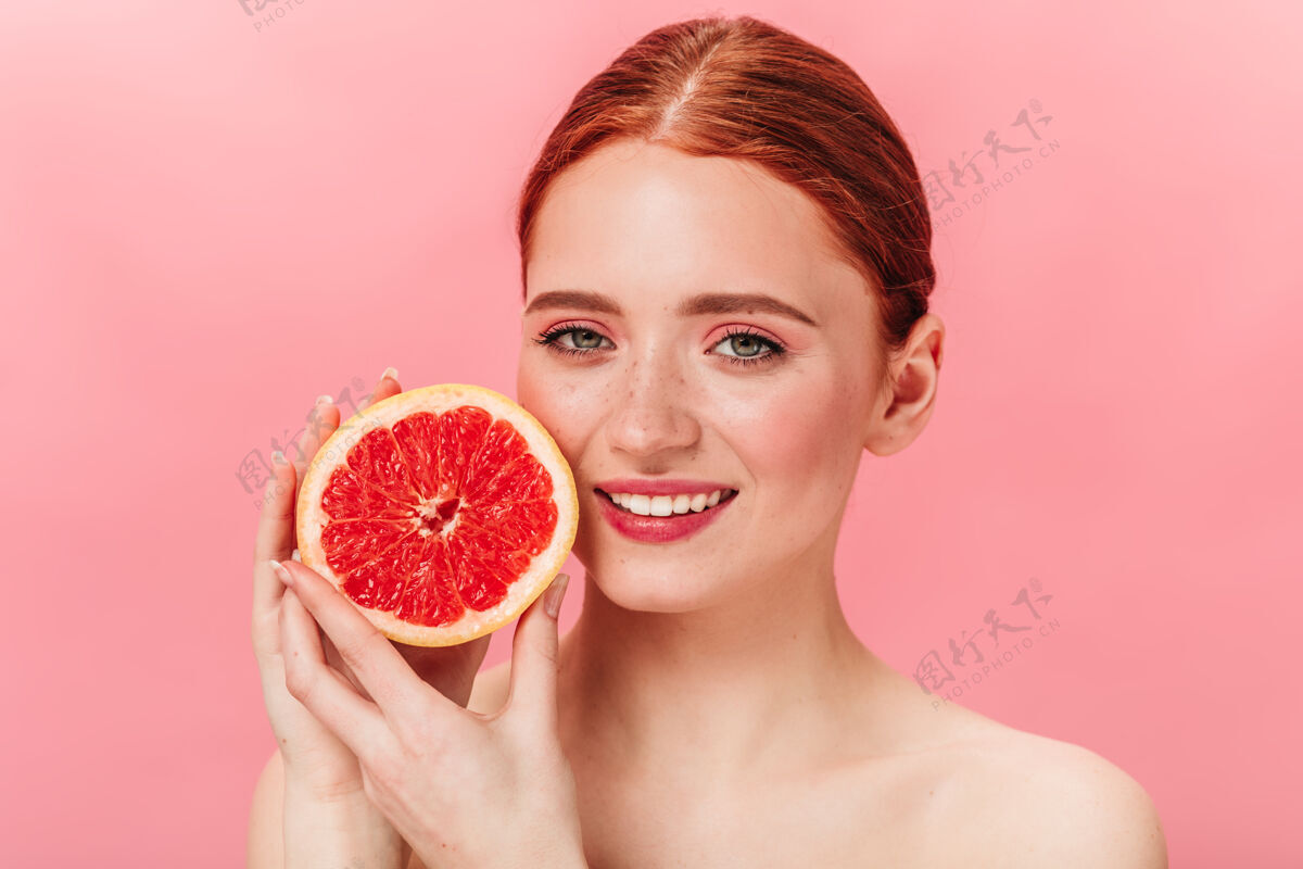 新鲜可爱的姜姑娘手里拿着多汁的葡萄柚漂亮的年轻女子在粉红色的背景上表达着幸福孤立的粉色女性微笑