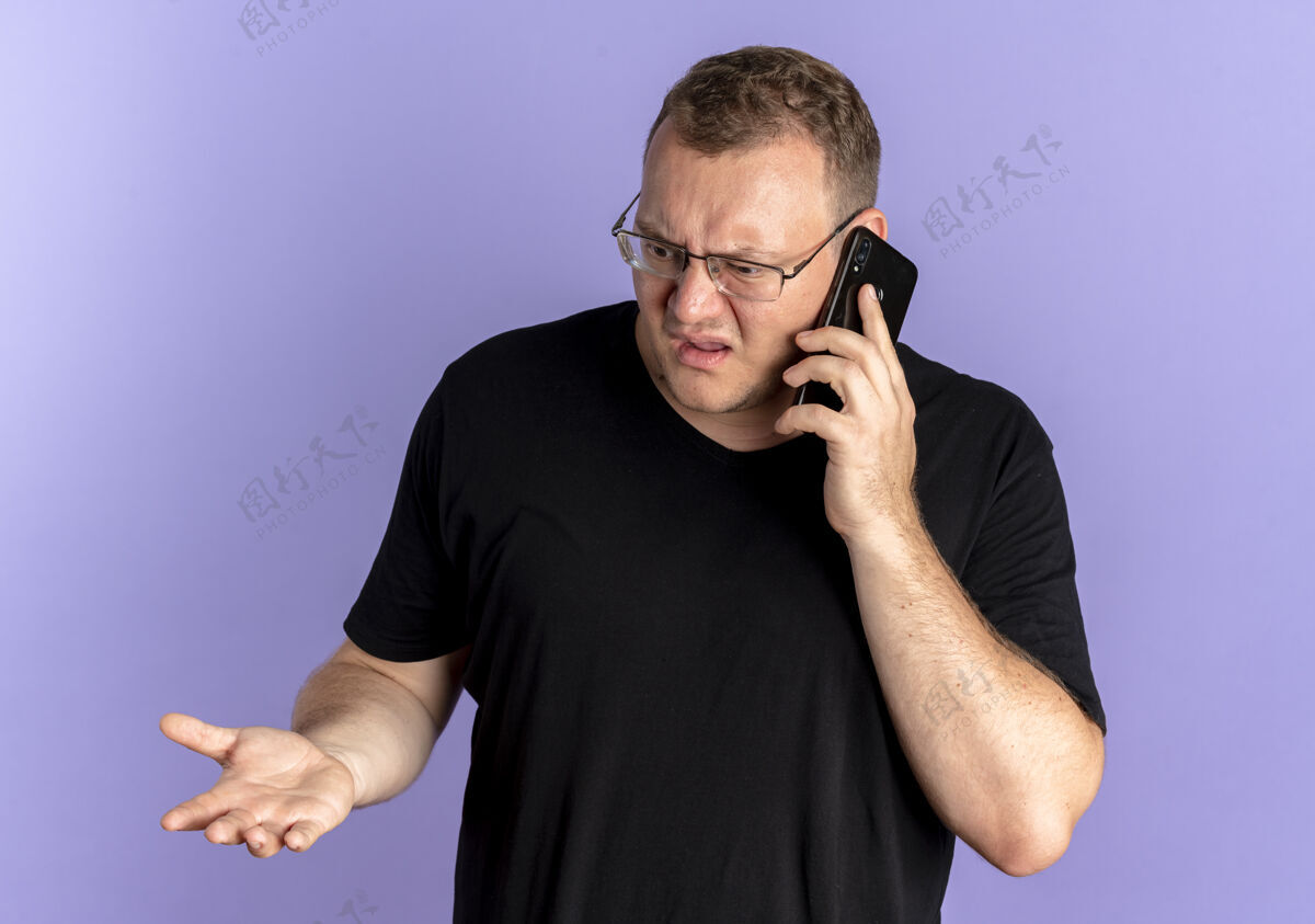 而戴眼镜的超重男子身穿黑色t恤 一边用蓝色手机交谈 一边显得困惑和不快电话黑色蓝色