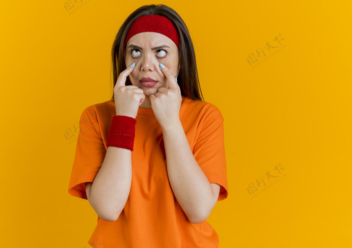 下戴着头带和腕带的年轻运动女性 拉着眼睑 交叉着眼睛向上看橙色盖腕带