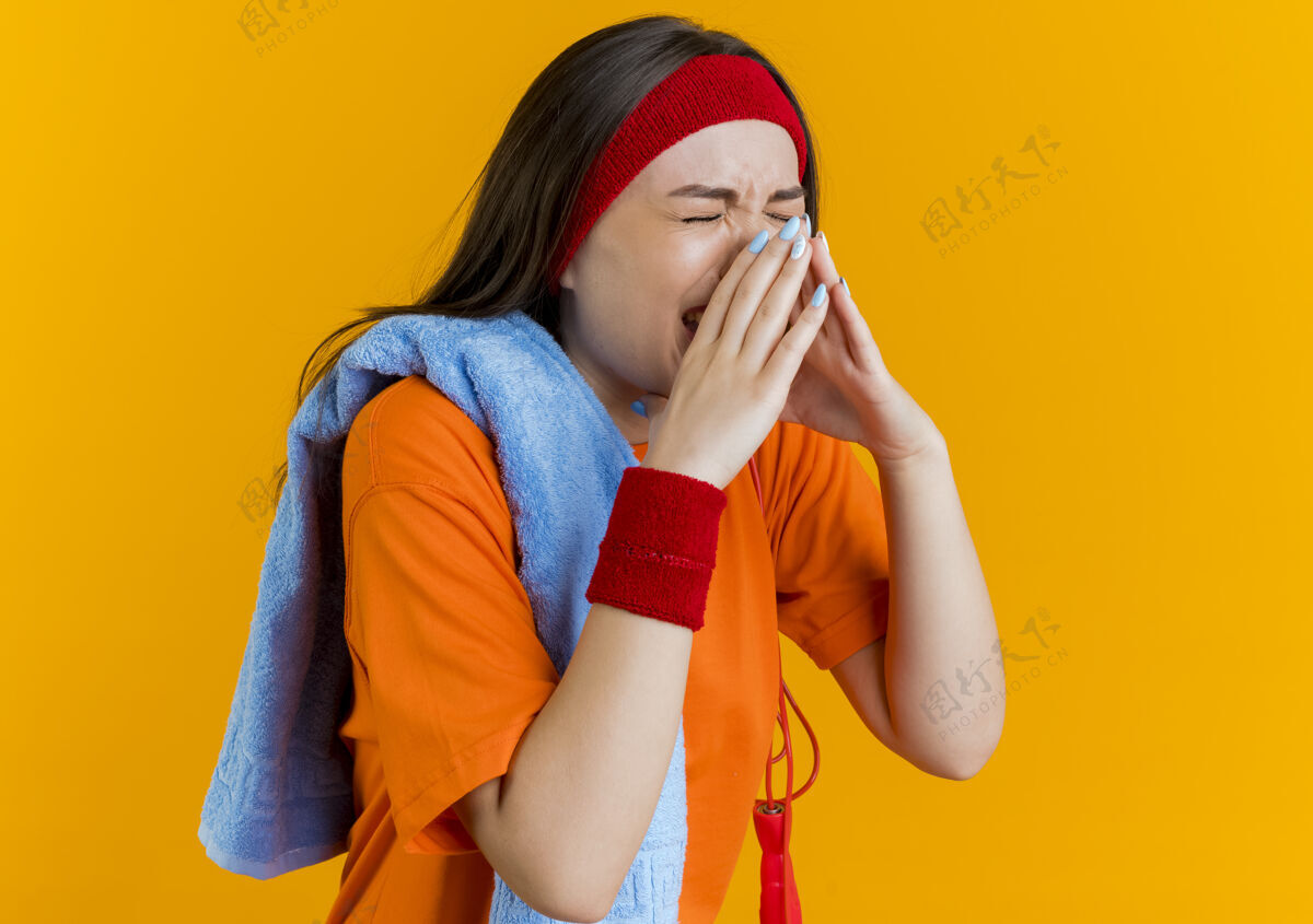 肩疼痛的年轻运动妇女戴着头带和手环与毛巾和跳绳在肩上抱着鼻子闭着眼睛腕带疼痛运动