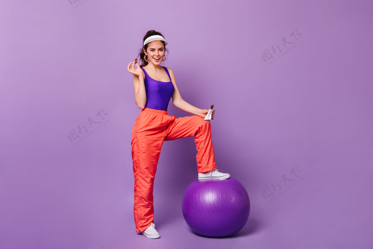 活动心情很好的美女把脚放在fitball上 在紫色的墙上摆出巧克力的姿势80年代风格姿势身材