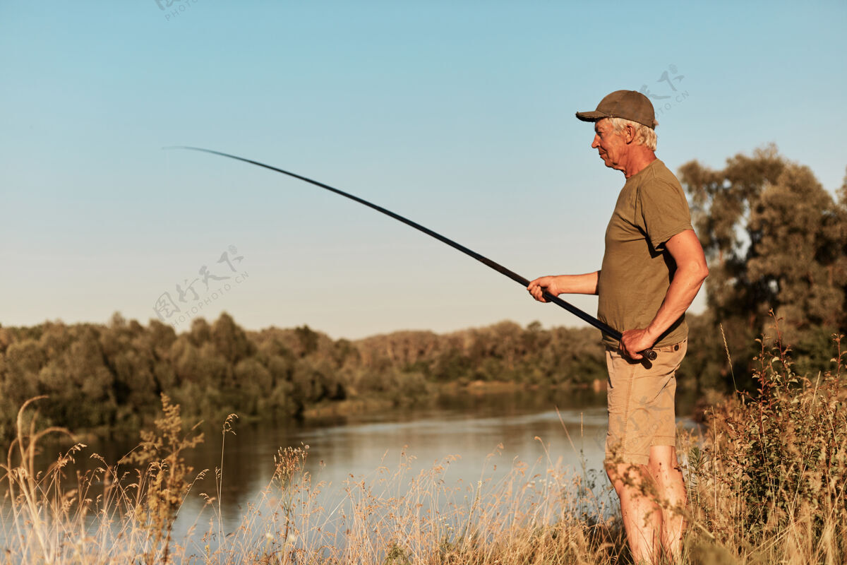运动渔夫站在湖畔或河畔望着手中的鱼竿 在夕阳下垂钓 在美丽的大自然中 穿着绿色的t恤和裤子湖休闲海岸