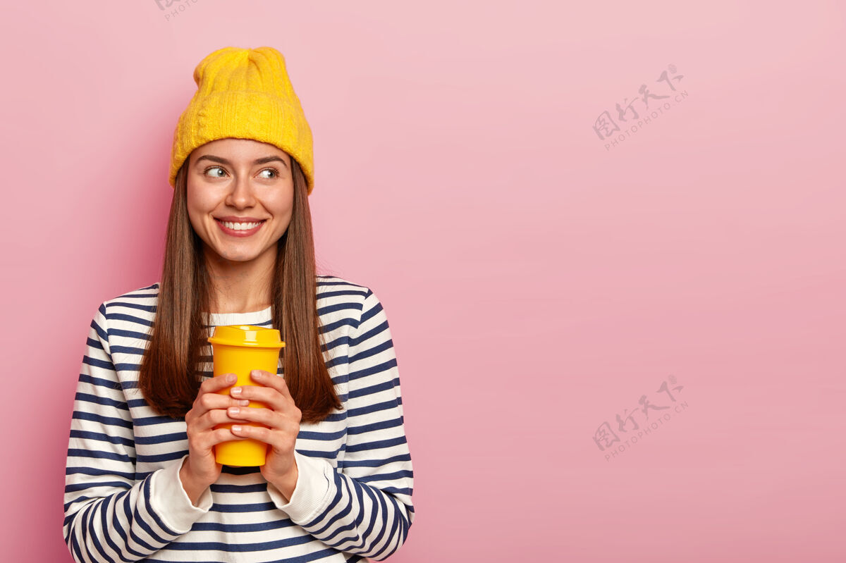 年轻人快乐的黑发女人的肖像拿着外卖咖啡 戴着黄色帽子和休闲条纹套头衫 微笑着看着旁边 隔着粉色的墙条纹咖啡请