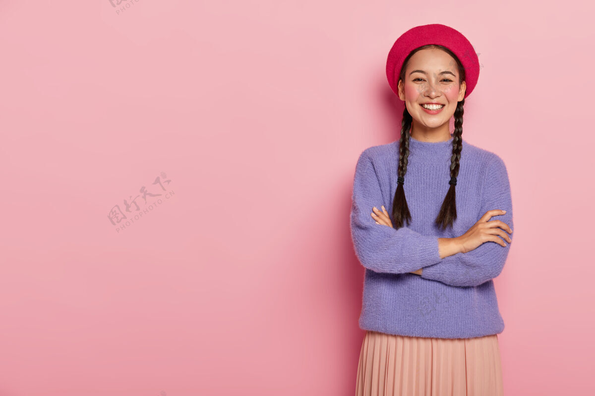 情感东方容貌的快乐女性肖像 双手交叉放在胸前 头戴红色贝雷帽 身穿紫色毛衣和裙子 对着粉色的墙壁摆姿势 看起来很热情时尚满意微笑