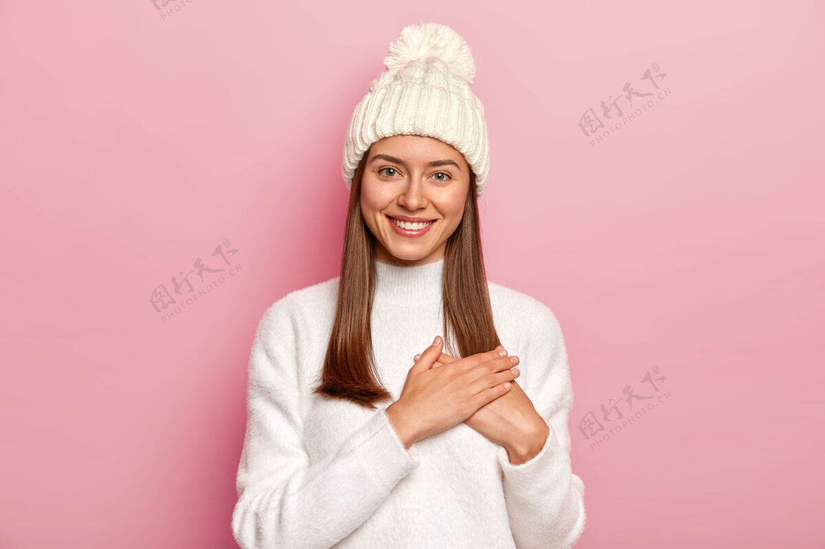 人类满意可爱的直发女人 从男朋友那里得到温暖的话语 做出感恩的手势 穿着舒适的白色毛衣和帽子 隔着粉色的墙黑发高兴手势