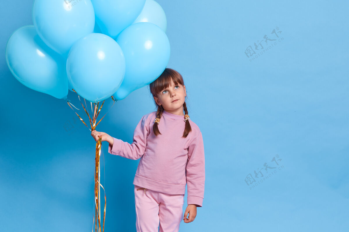 思想可爱的欧洲女孩儿戴着玫瑰结 留着小辫子的孩子面带深思熟虑的表情 梦想着一些愉快的事情 抱着一堆氦气球 靠着蓝色的墙壁气球沉思女孩