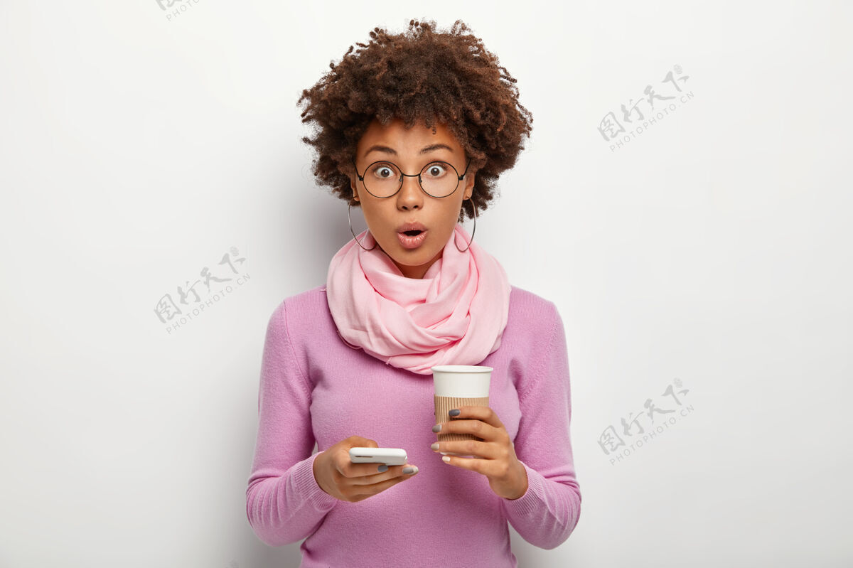 饮料秀发清爽的惊艳美女查看新闻 收到震惊的信息 拿着外卖咖啡 不敢相信什么 戴着眼镜和紫色毛衣 在室内摆姿势忙碌惊喜印象