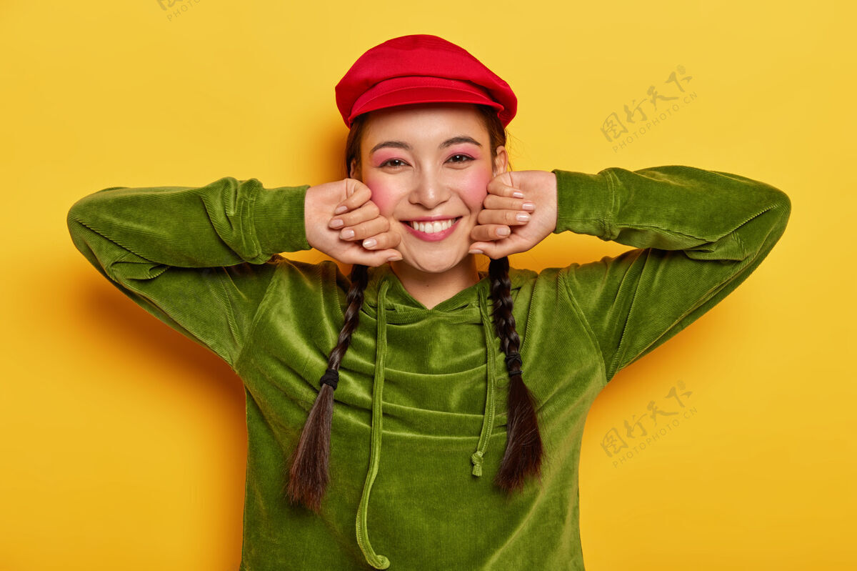 姿势积极的女孩抚摸脸颊 双手侧放 微笑愉快 戴着红色贝雷帽和绿色天鹅绒运动衫胭脂亚洲人化妆