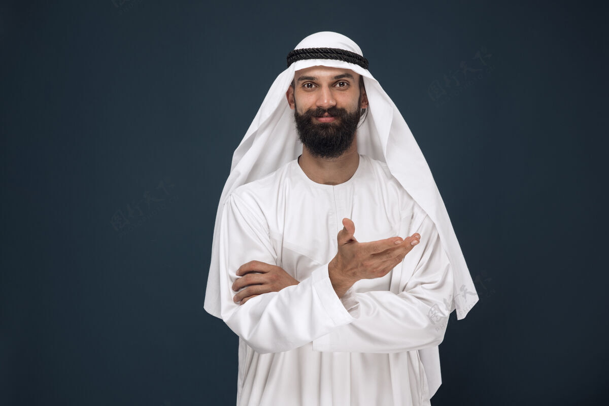面部深蓝色墙上的半身阿拉伯沙特商人肖像年轻的男模微笑着指着商业 金融 面部表情 人类情感的概念惊喜发呆胡须
