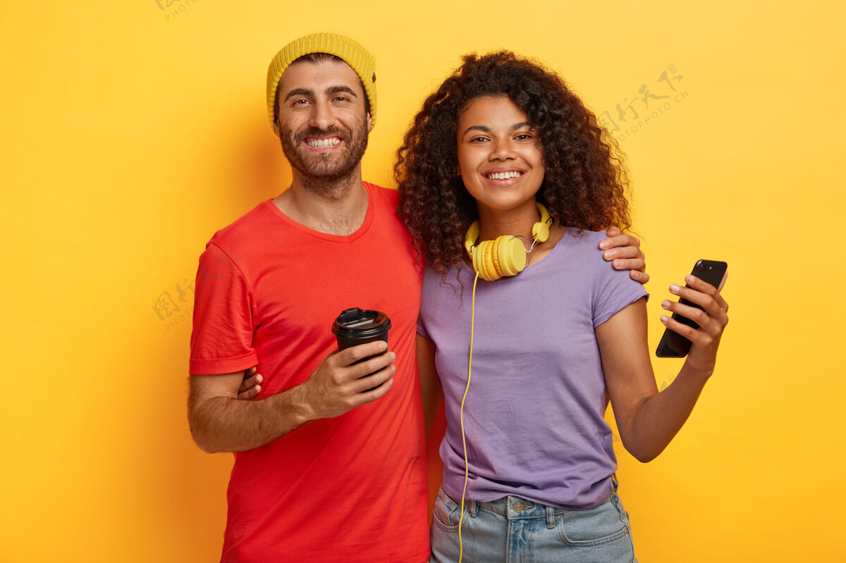电话情侣们在一起消磨空闲时间 喝咖啡 使用现代手机进行在线交流 穿着t恤 在黄色背景下紧紧地站在一起女朋友乐趣小工具
