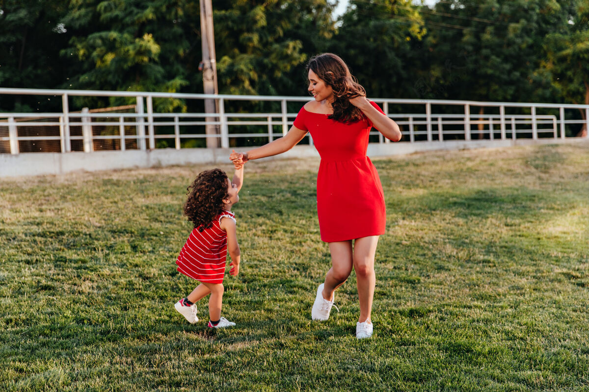 游戏快乐的红衣女子和她的女儿在草坪上跳舞在公园里玩的黑发女孩和小孩的户外全长肖像游戏草地拥抱