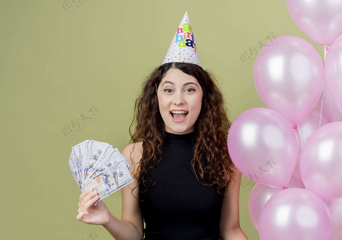 站立一个年轻漂亮的女人 卷发 戴着节日帽 手里拿着气球 快乐而兴奋地展示着现金生日聚会的概念 站在光墙上光年轻空气