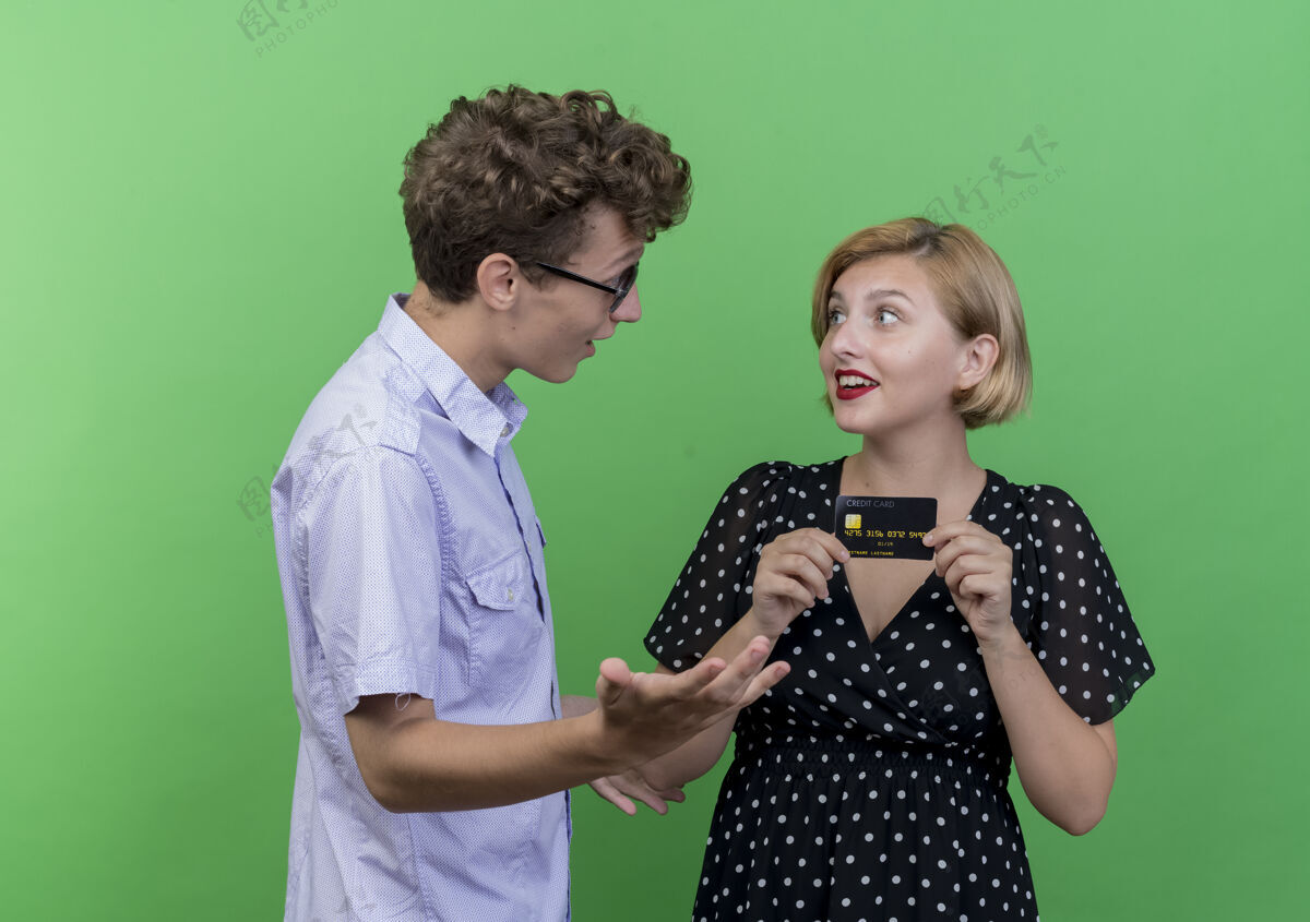 信用年轻漂亮的夫妇惊讶地看着那个拿着信用卡站在绿墙上的快乐女友持有立场女朋友
