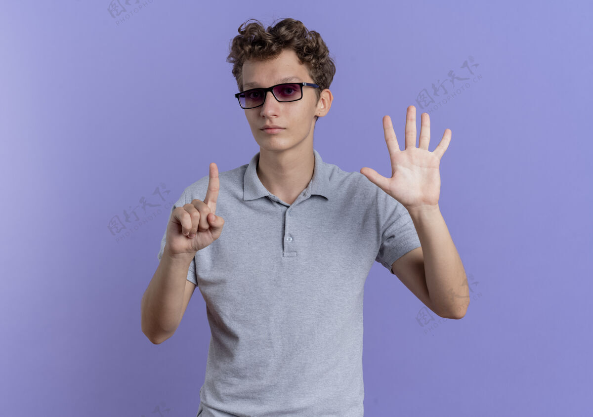 马球一个戴着黑眼镜的年轻人 穿着灰色马球衫 脸上露出丝线 手指向上 六号手指站在蓝色的墙上秀男人穿