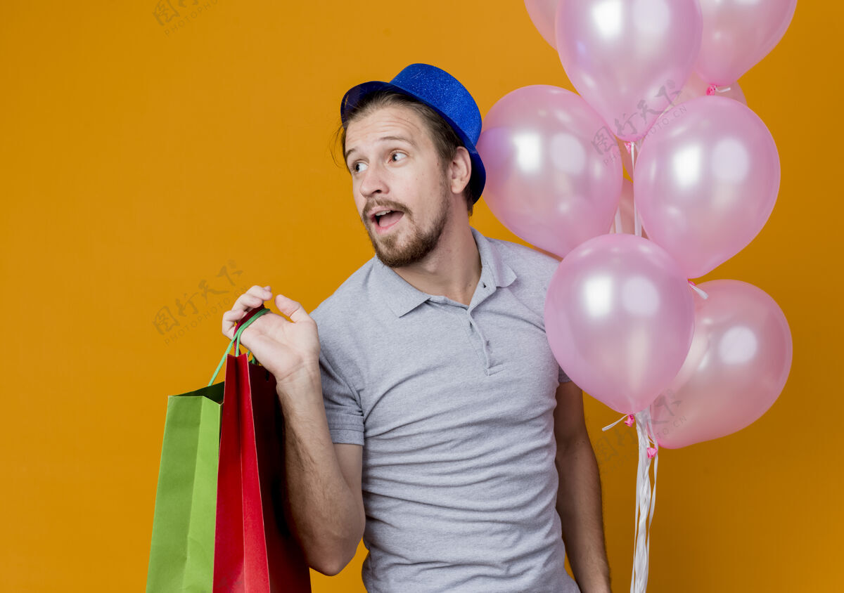 抱着一个戴着帽子的年轻人举着纸袋和气球站在橘色的墙上欢笑着庆祝生日兴奋男人站着