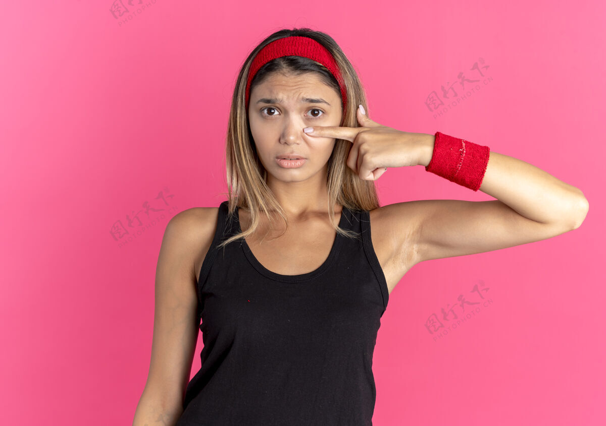 站身穿黑色运动服 头戴红头巾 手指抵着鼻子的年轻健身女孩站在粉红色的墙上 表情悲伤年轻表情运动装