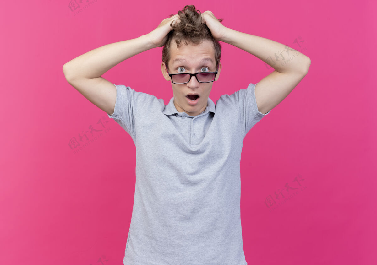 站一个戴着黑色眼镜 穿着灰色马球衫的沮丧的年轻人 站在粉红色的墙上 摸着头 拉着头发穿拉年轻