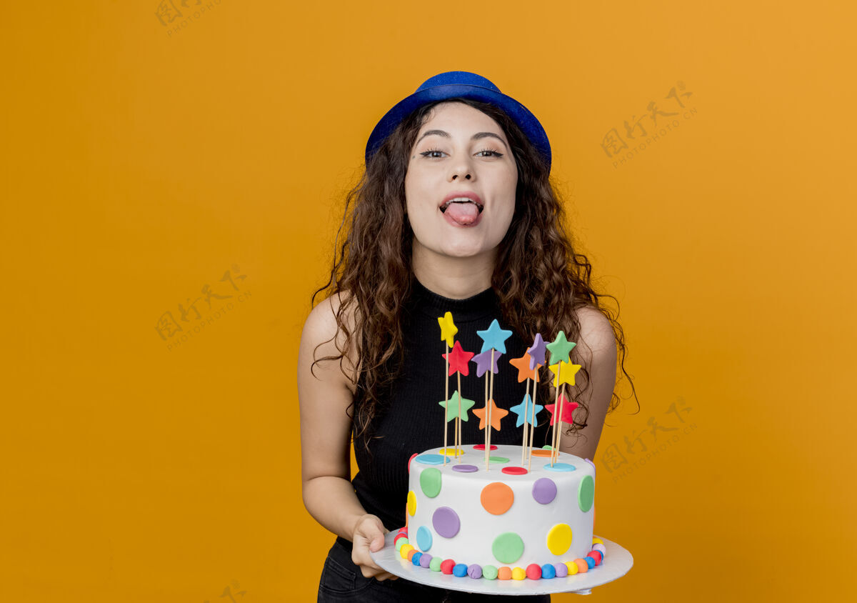 蛋糕一个年轻漂亮的女人 卷发 戴着节日礼帽 手里拿着生日蛋糕 开心地伸出舌头站在橙色的墙上年轻舌头女人