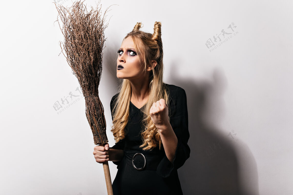 成人愤怒的年轻女巫拿着扫帚举起拳头一张金发碧眼的女孩带着浓妆 穿着万圣节服装的照片扫帚优雅魔术