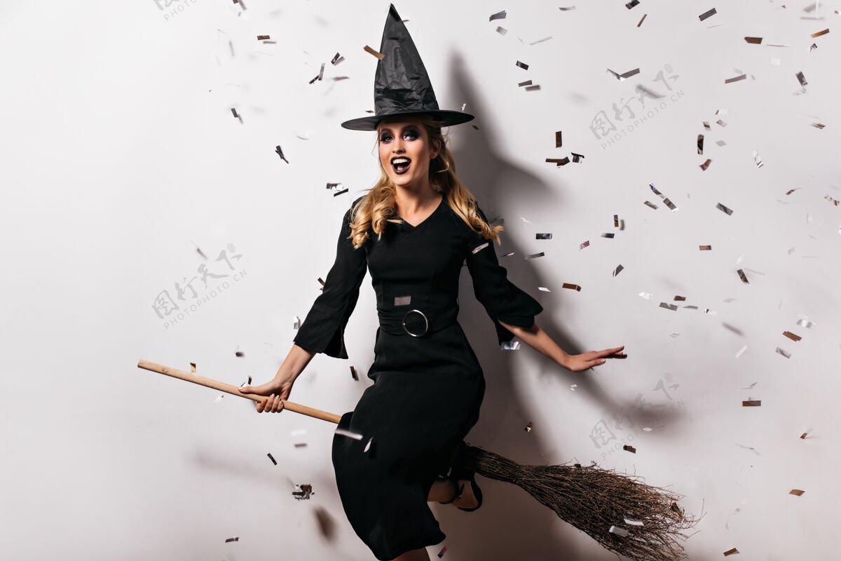 幽灵穿着黑色衣服 身材匀称的年轻女巫坐在扫帚上万圣节派对上 可爱的巫师戴着帽子 穿着长裙的室内镜头庆典微笑邪恶