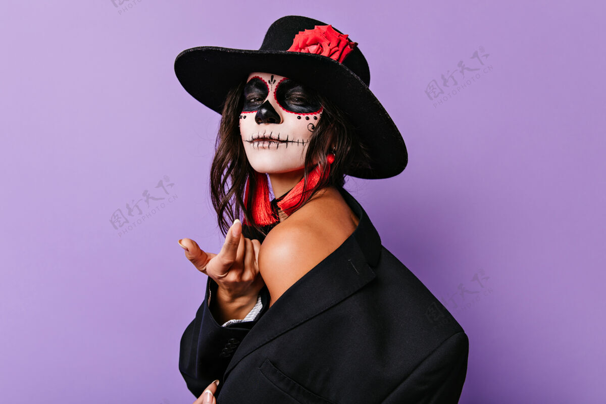 恐怖站在紫色墙上的优雅的年轻女子 身着黑色罩衫 带着万圣节彩妆的黑发女孩 无忧无虑地微笑着事件严峻死亡之日