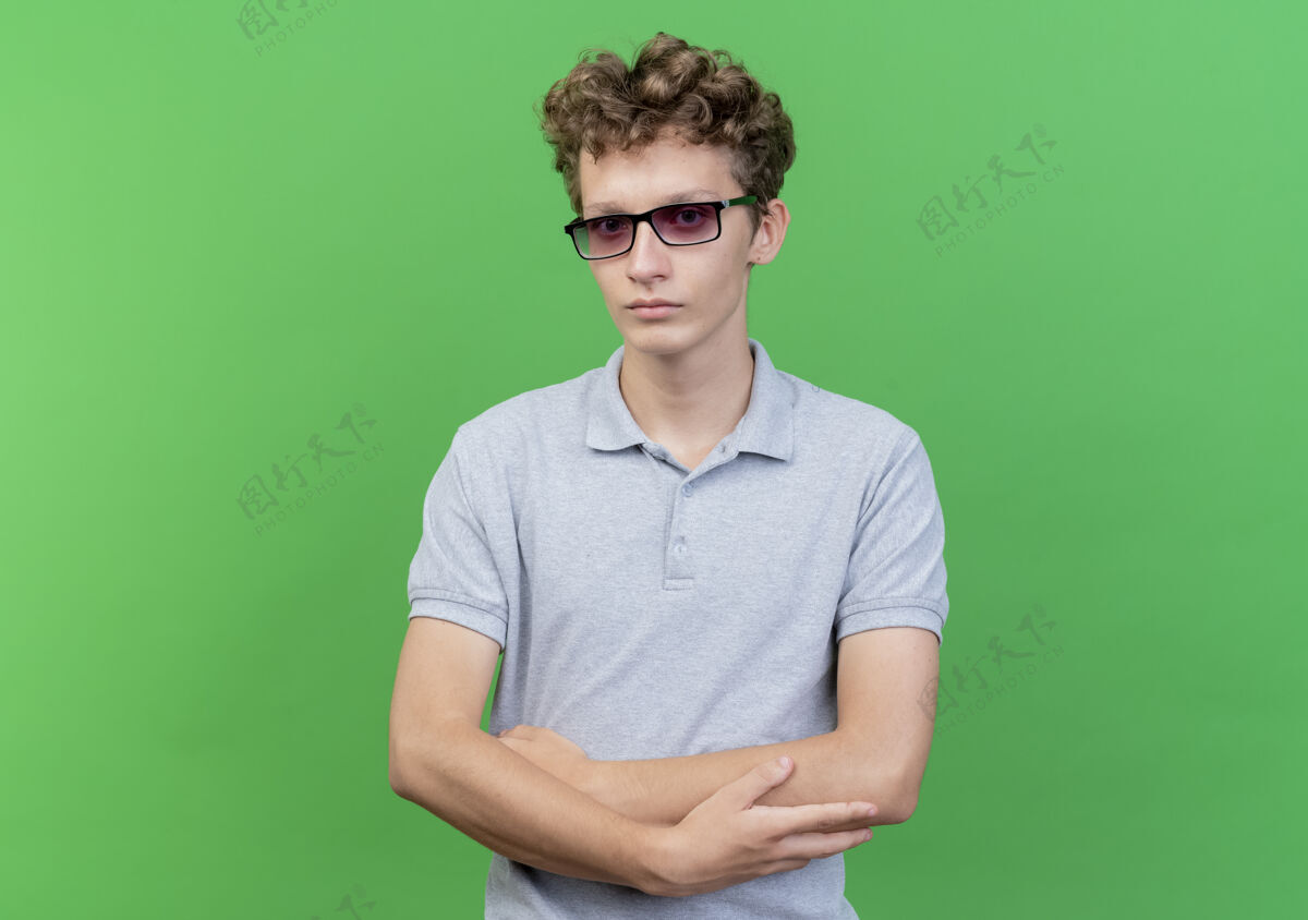 交叉戴着黑眼镜的年轻人穿着灰色马球衫 严肃的脸交叉着双手站在绿色的墙上站眼镜严重