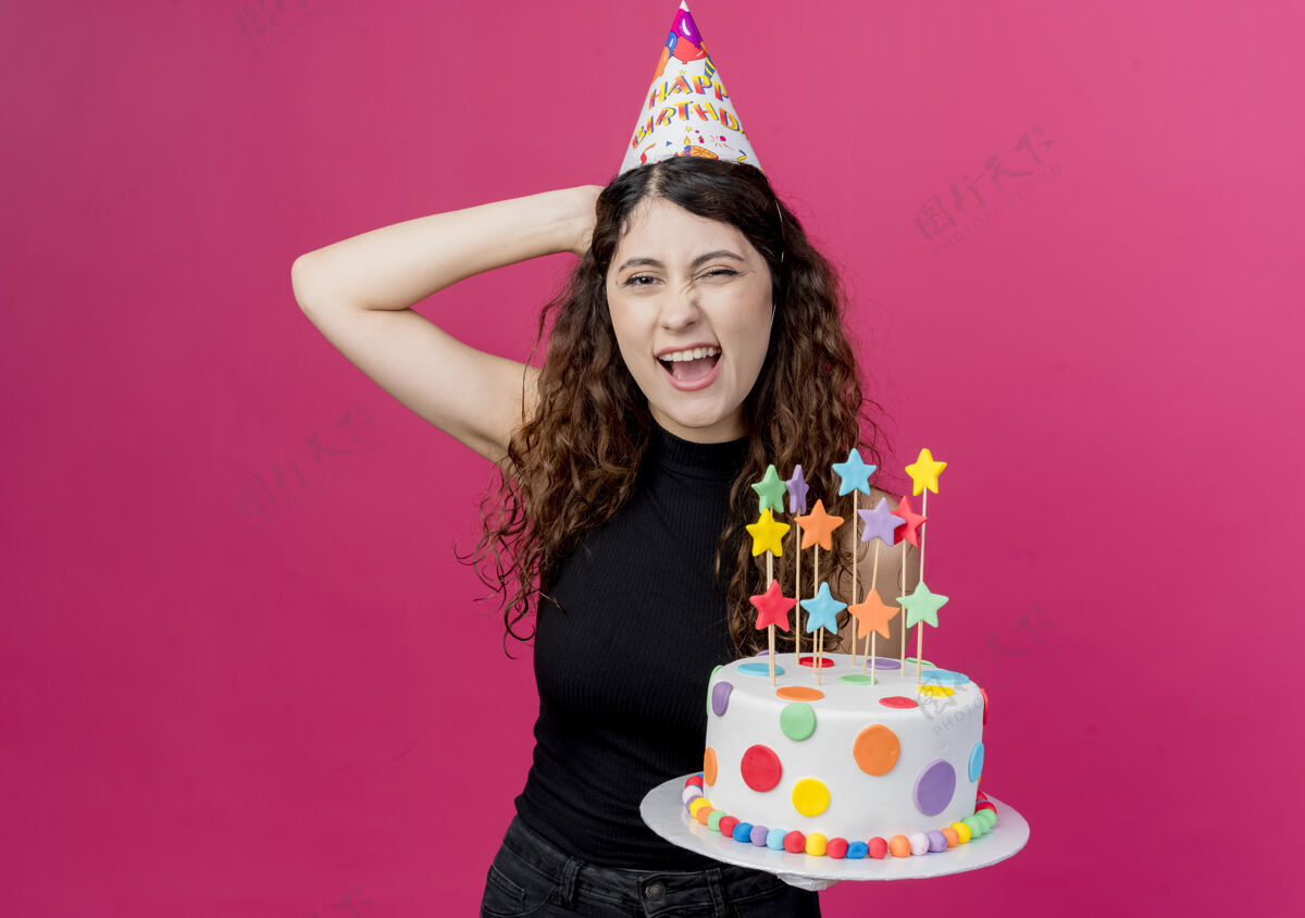 看着一位年轻漂亮的女士 卷发 戴着节日帽 手里拿着生日蛋糕 面带微笑 站在粉色的墙上 看起来很开心 很快乐欢呼站着年轻