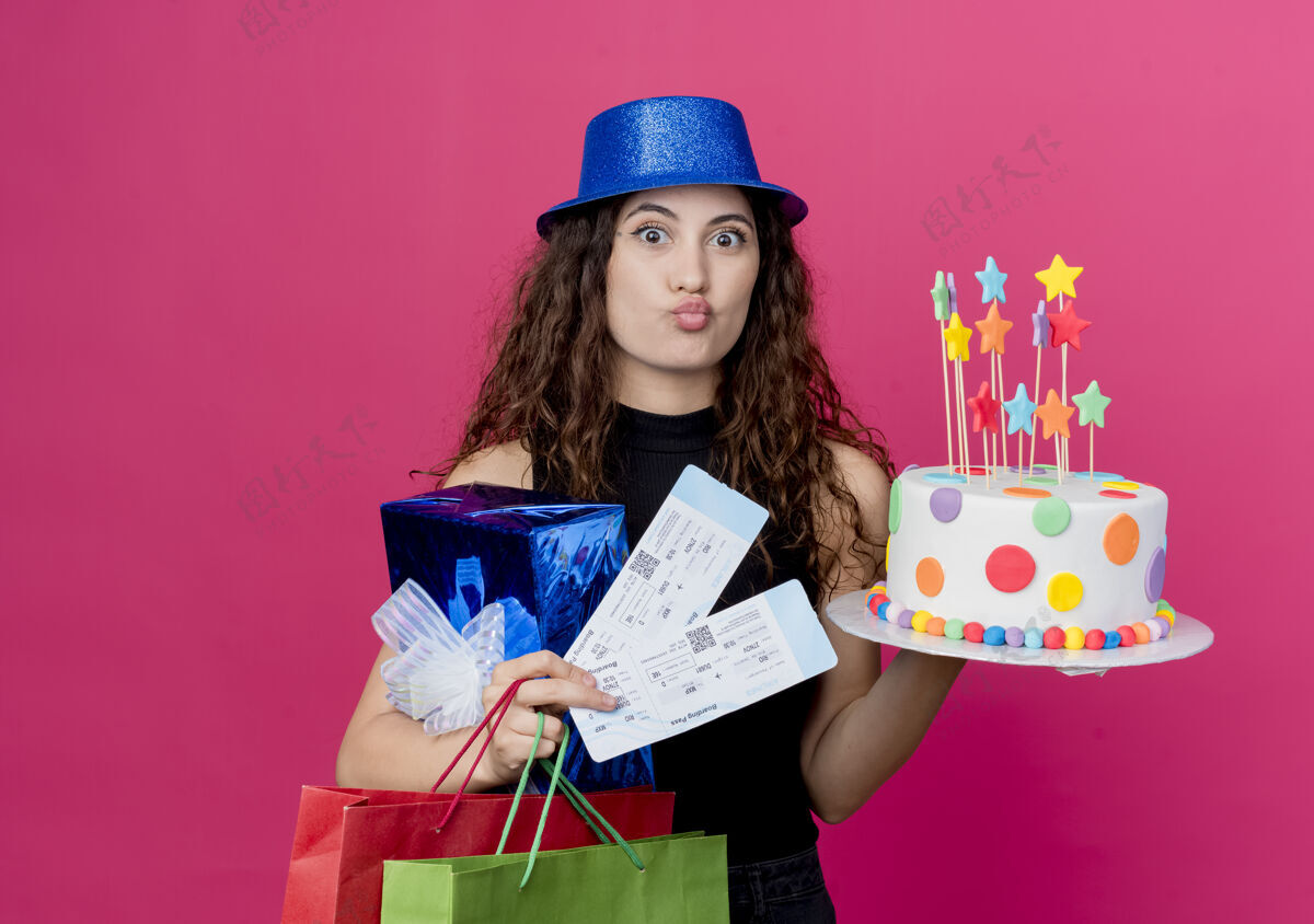 飞机一位年轻漂亮的女士 卷发 戴着节日礼帽 手里拿着生日蛋糕礼盒和机票 站在粉色的墙上 快乐而积极的生日派对理念拿着正面蛋糕