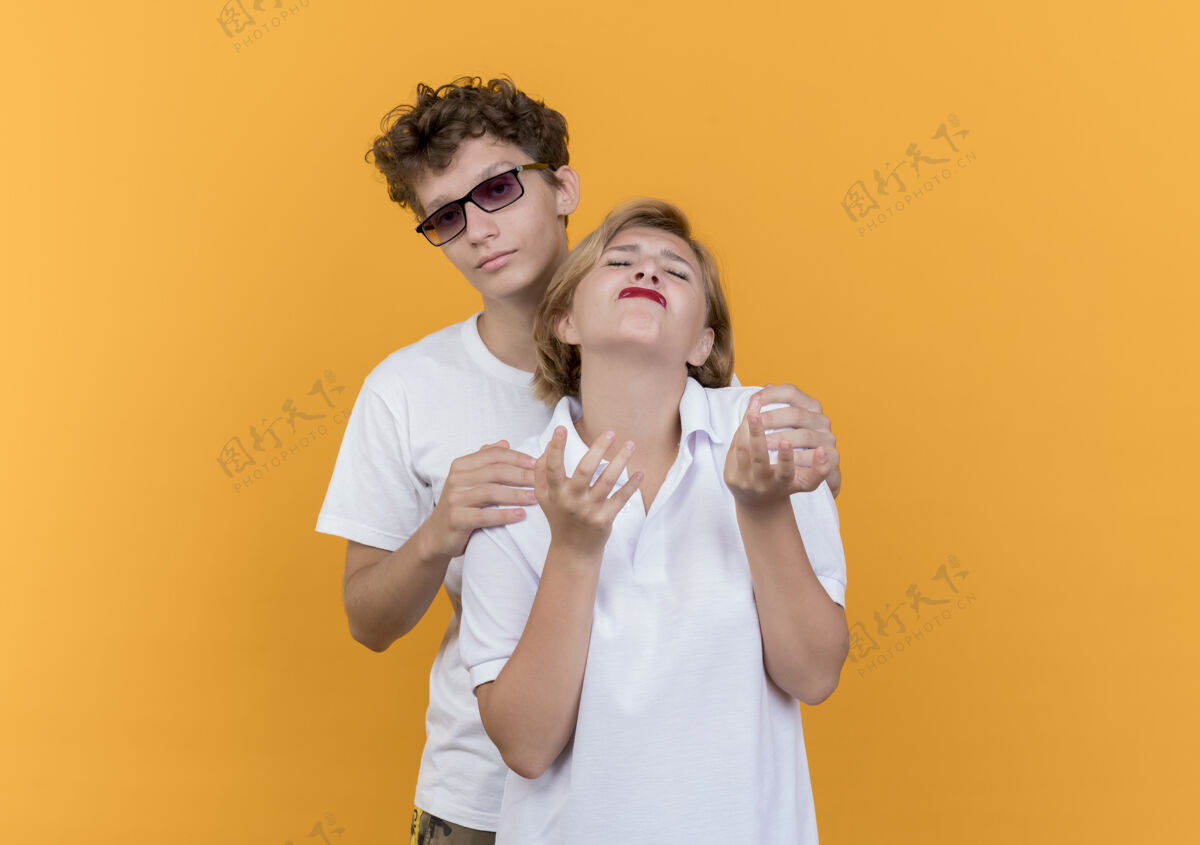站立一对年轻的情侣站在橘色的墙上 试图拥抱他不高兴和不安的女友年轻男人尝试