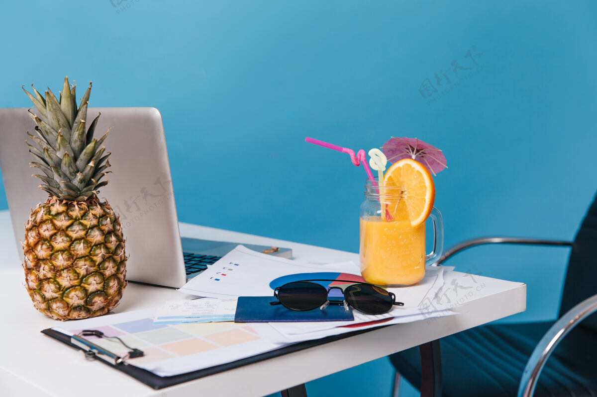 鸡尾酒文件 太阳镜 鸡尾酒 菠萝和笔记本电脑的快照放在蓝色空间的桌子上官方办公桌帽子