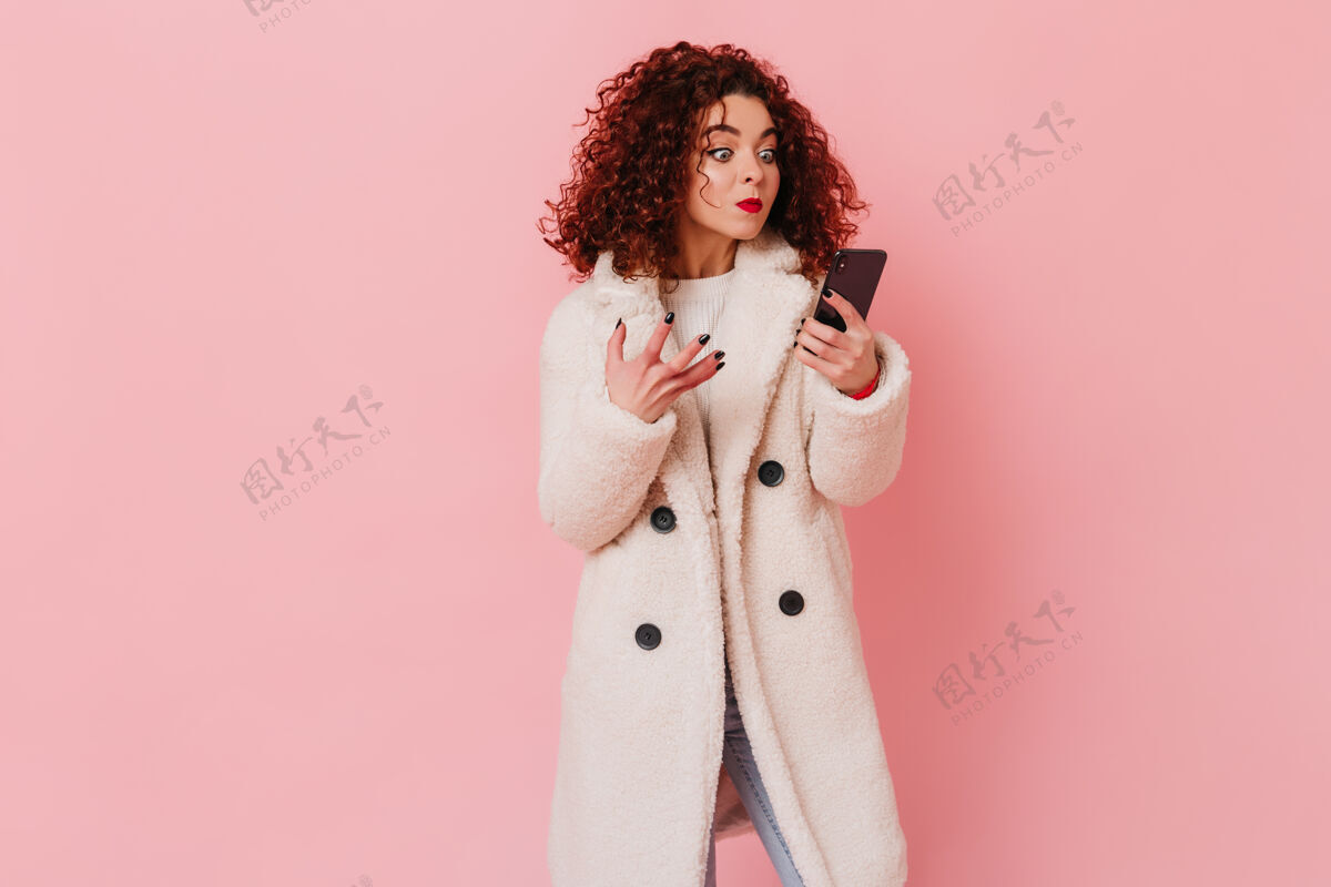积极不满意的女孩看着手机屏幕生气粉红色的空间里有一个卷曲的女人的肖像 嘴唇明亮 穿着白色外套时尚莫比尔发型