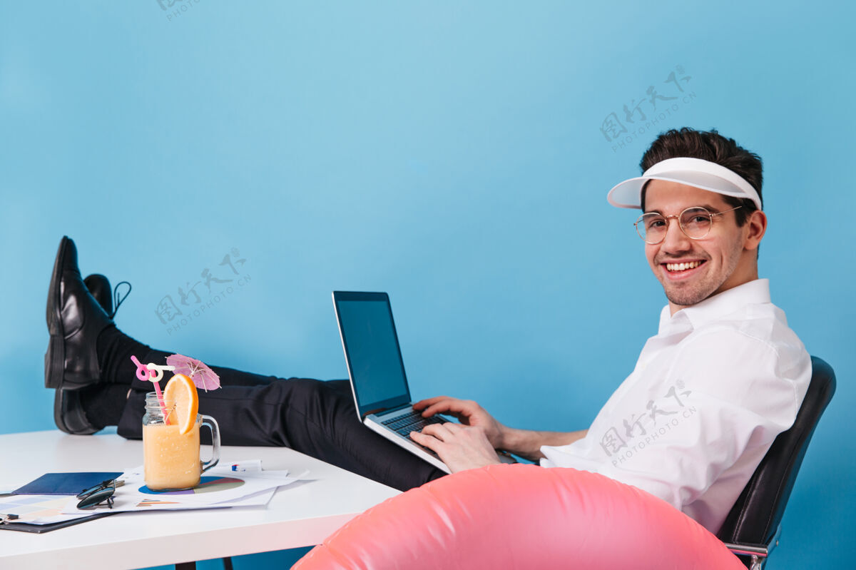 正式戴着眼镜和帽子的男人在一个孤立的空间里工作男人拿着笔记本电脑 鸡尾酒和充气圈摆姿势文件成功员工
