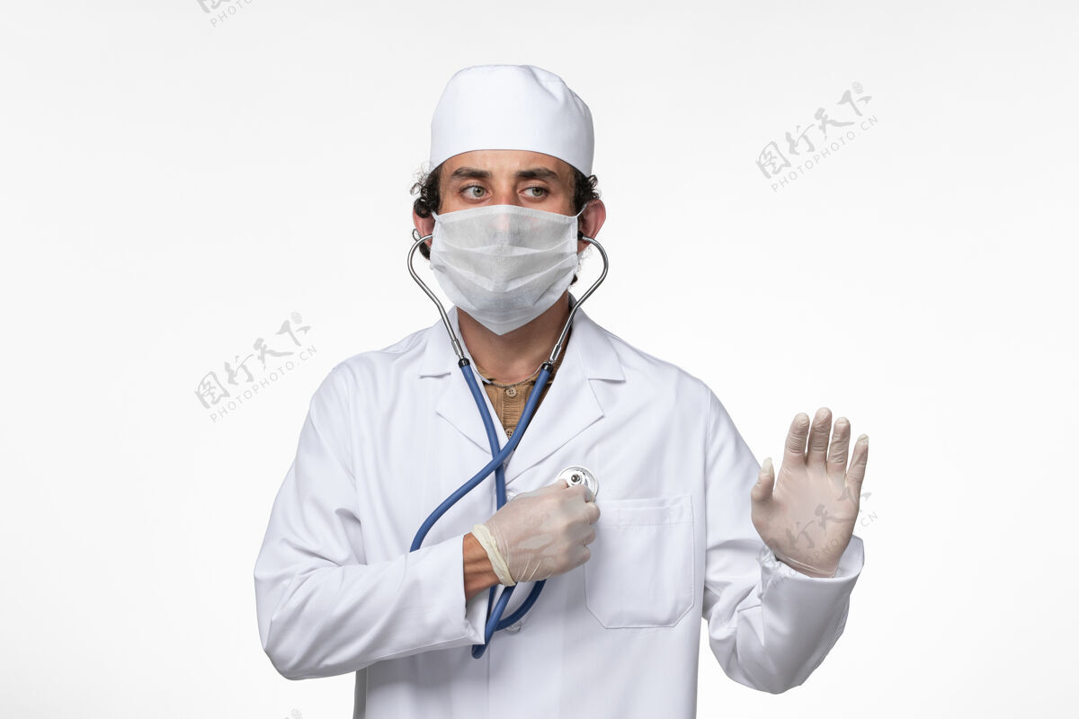 冠状病毒防护正面图：男医生穿着医疗服 戴着口罩以防病毒感染-使用听诊器对白墙病病毒进行病毒感染-大流行视图病毒使用