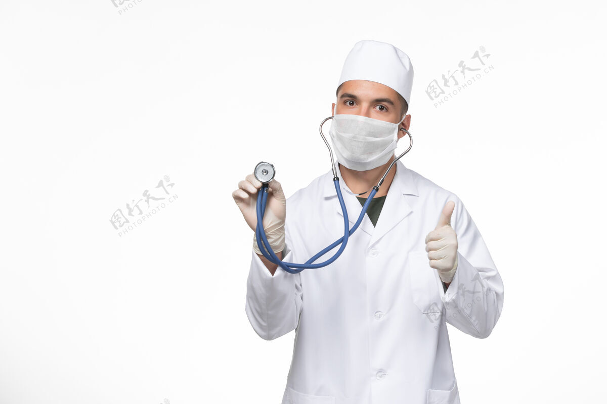 病毒正面图：男医生穿着医疗服 戴着防冠状病毒口罩 在白墙上用听诊器观察冠状病毒疾病大流行正面医疗视图