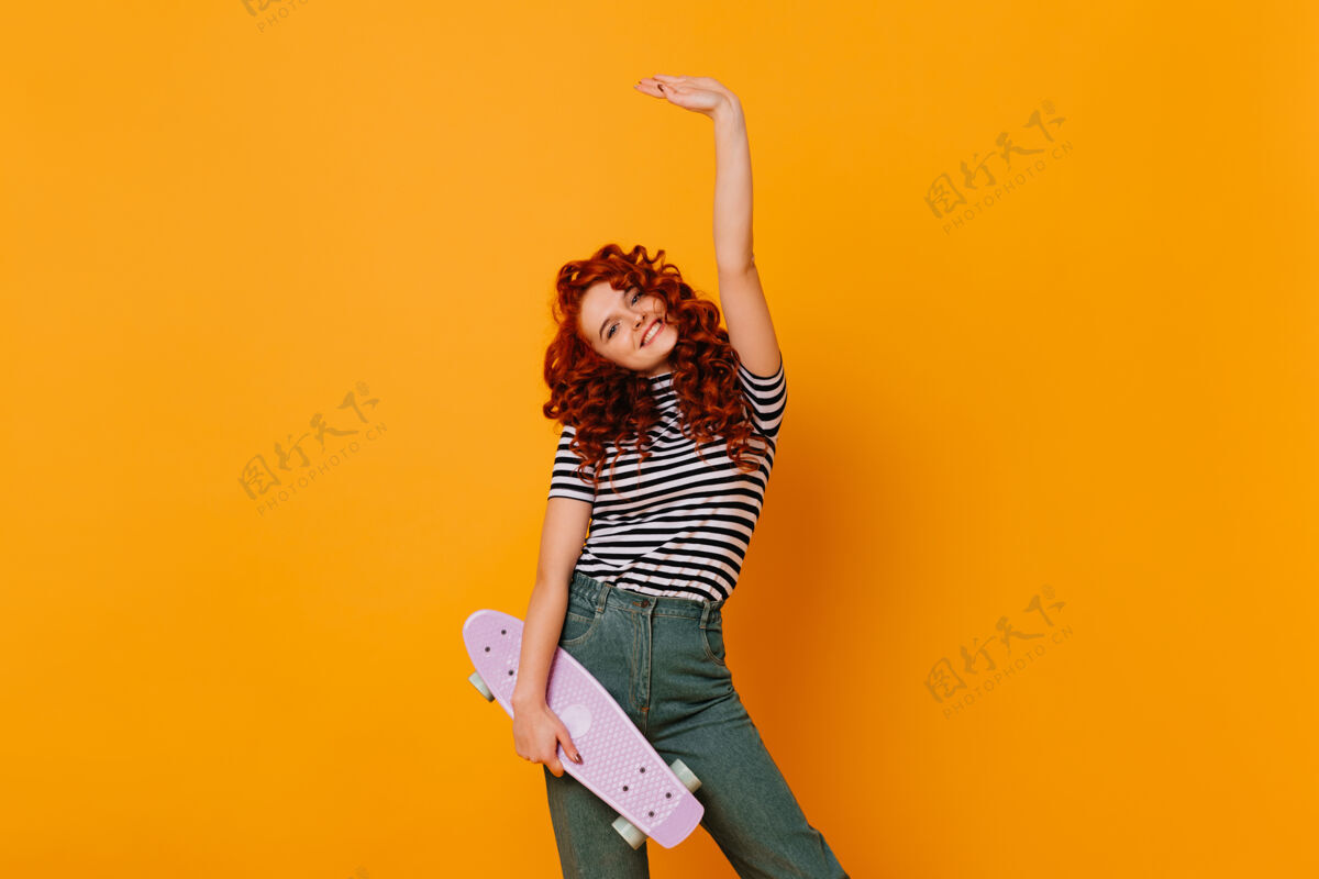 牛仔一头红色卷发的快乐女孩举起手来 在橙色的空间里和溜冰者合影时尚表情兴奋