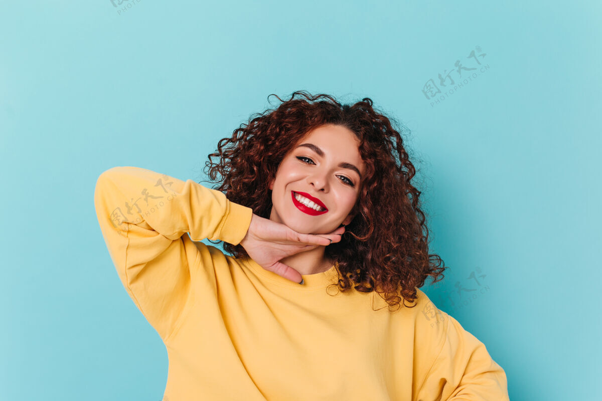 自信蓝眼睛红唇女孩的特写照片穿着黄色毛衣的卷发女人抚摸着下巴 在蓝色空间里摆姿势头发高兴红唇