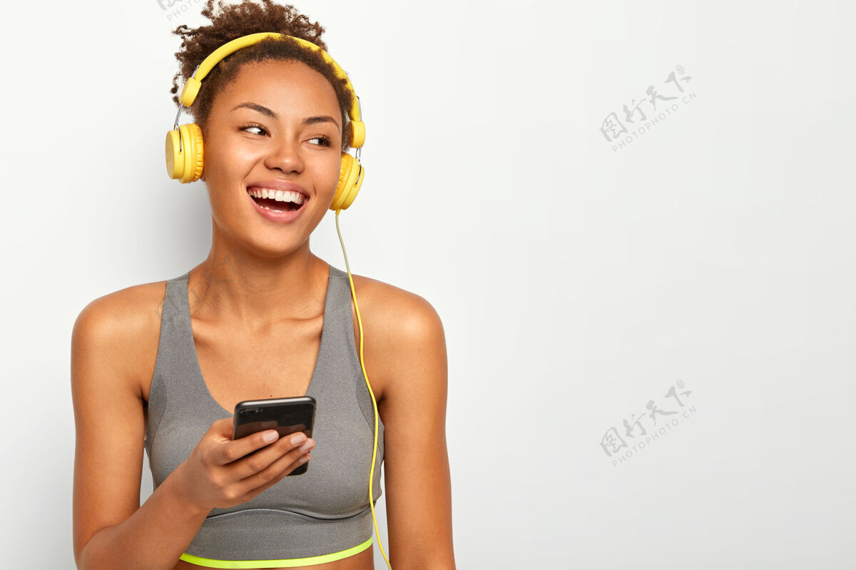 身体喜出望外的黑皮肤女运动员笑从愉快的情绪 持有现代细胞 转身离开 穿着运动服 有积极的训练与音乐高兴非洲电话