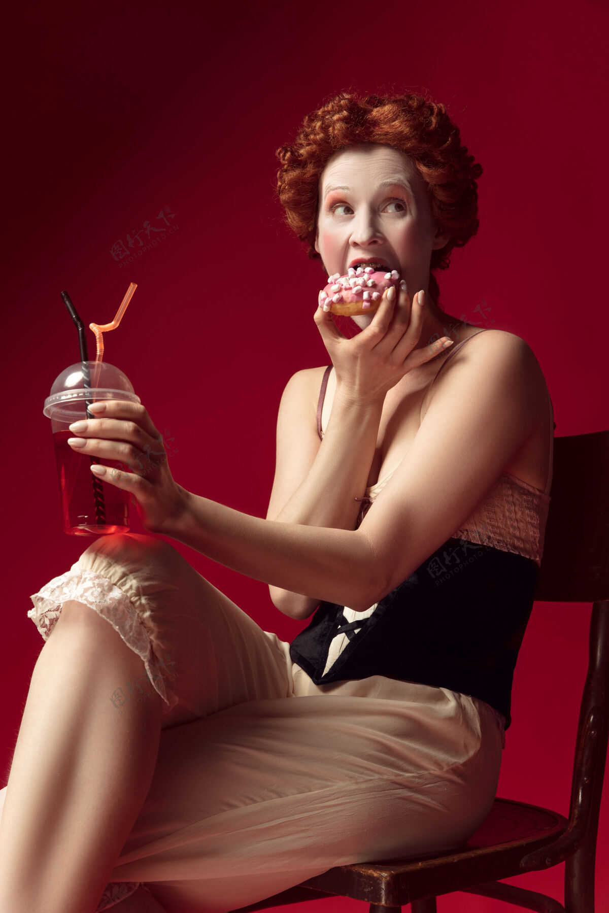 骄傲中世纪的红发女公爵 身穿黑色紧身衣和睡衣 坐在红色空间的椅子上 喝着饮料和甜甜圈礼服文艺复兴情感