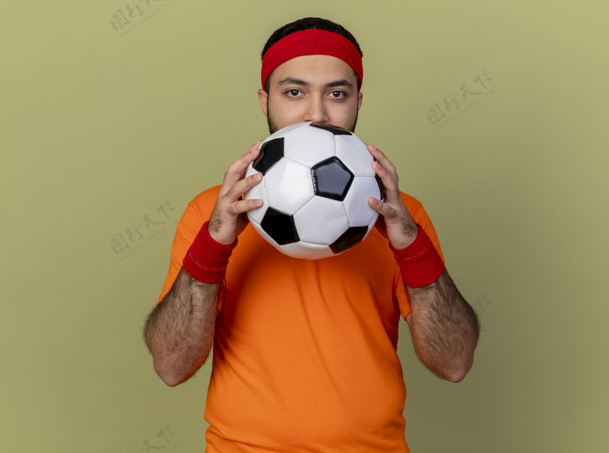 运动年轻的运动型男子看着相机戴着头带和手环拿着球孤立在橄榄绿年轻相机球