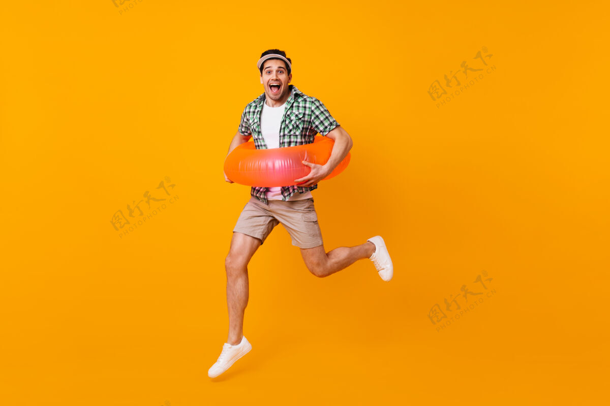 衣服棕色眼睛的深色男人穿着米色短裤和绿色t恤 在橙色的空间里用充气圈跳跃鸡尾酒水果行李