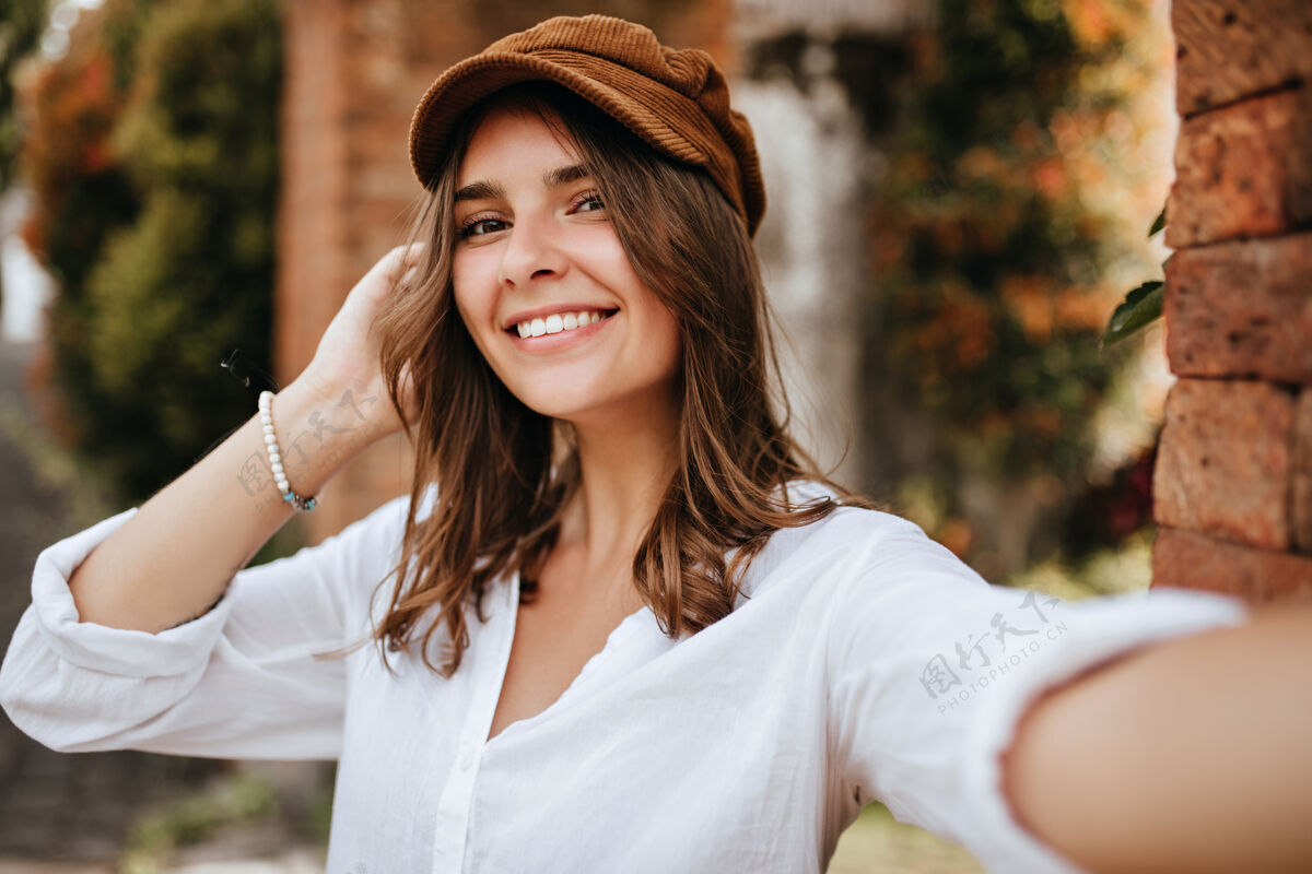 深金发棕色眼睛的女孩 头戴天鹅绒帽子 穿着白色上衣 在砖墙和树木的空间里自拍裤子街道砖房