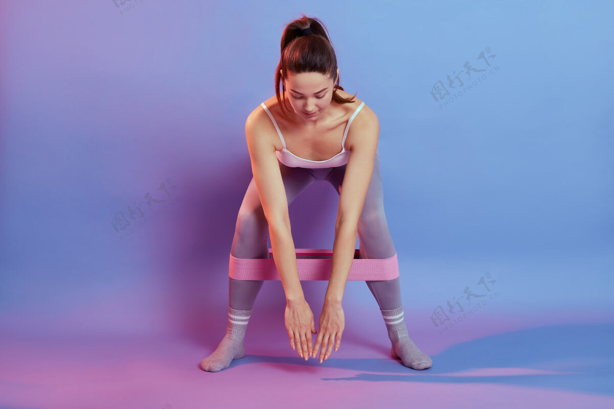 肌肉穿着运动服的女孩在腿上系上阻力带 将身体靠在彩色背景上 女性下蹲 俯视 在室内锻炼的全长照片健身漂亮姿势