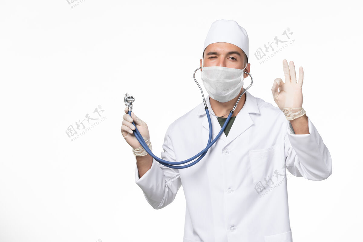 听诊器正面图男医生穿着医疗服 戴着口罩因冠状病毒-用听诊器眨眼白墙冠状病毒-大流行病毒病实验室外套口罩男性