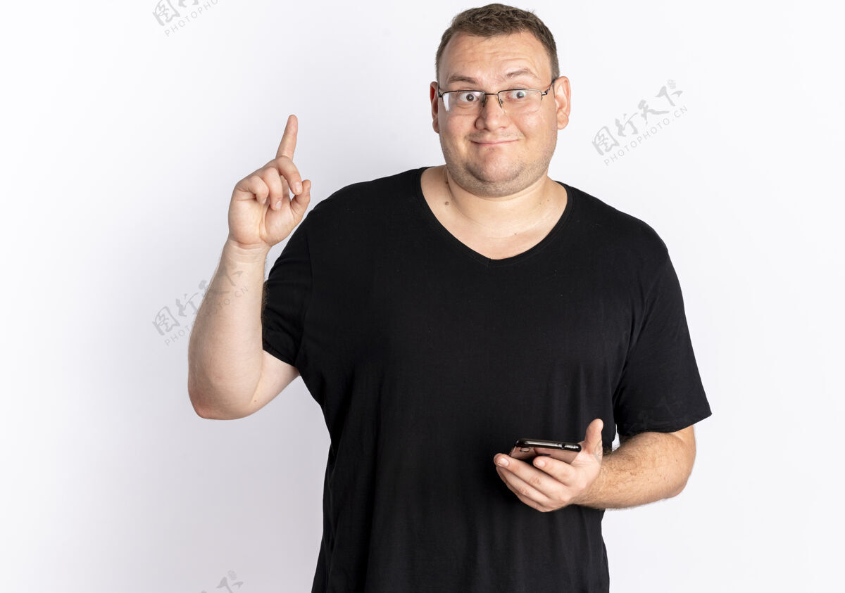 展示戴眼镜的超重男子身穿黑色t恤手持智能手机显示食指有新想法站在白墙上站着拿着男人