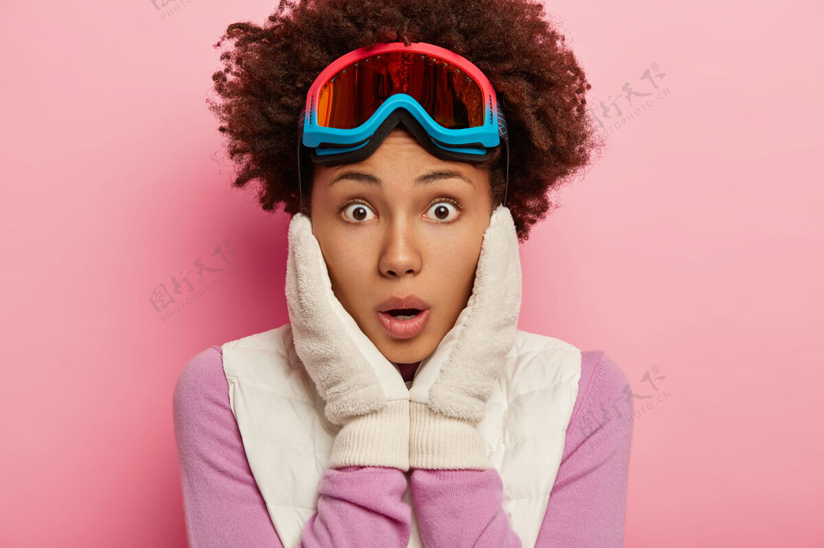 运动照片中惊呆了的情绪卷曲的女性在镜头前表情震惊 双手放在脸颊上 戴着滑雪眼镜滑雪 隔离在玫瑰色的工作室墙上惊呆了非洲头像
