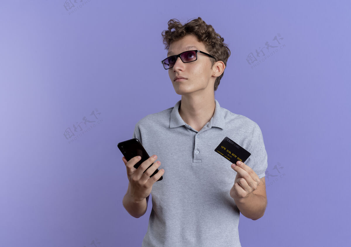 男人一个戴着黑眼镜 穿着灰色马球衫 拿着智能手机和信用卡的年轻人站在蓝色的墙上困惑地看着我穿拼图手机