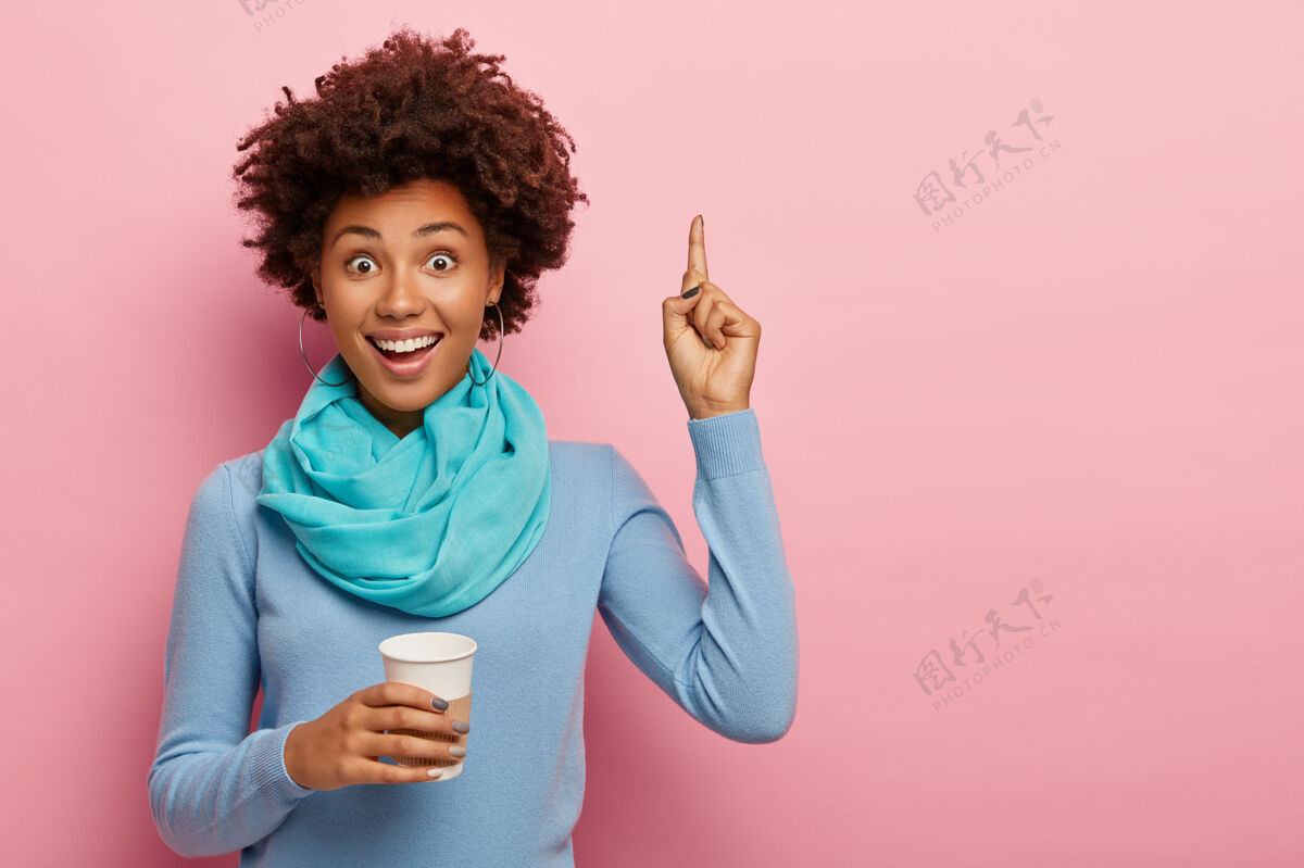 站着快乐的非洲裔美国妇女的照片指向上方 向上展示一些东西 有咖啡休息时间 拿着纸一次性杯子 穿着蓝色套头衫 有快乐的表情 在粉色背景上摆姿势室内兴奋外卖