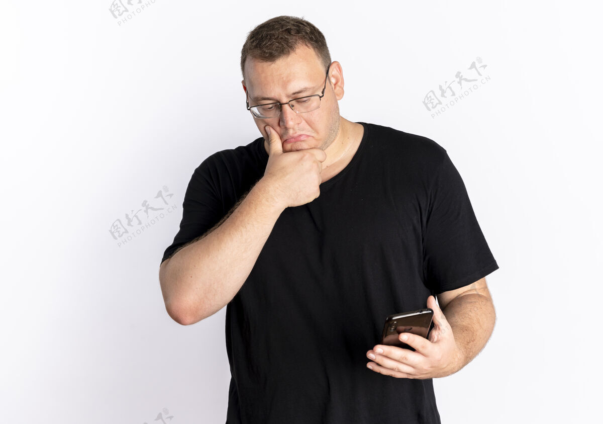 穿一个戴眼镜 身穿黑色t恤的超重男子站在白色的墙上困惑地看着他的智能手机屏幕看眼镜站