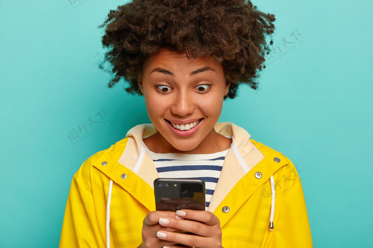 手持快乐的时髦女孩 带着惊奇的表情 读着愉快的短信 喜欢聊天 穿着黄色雨衣隔离在蓝色的墙上牙关秋天非洲