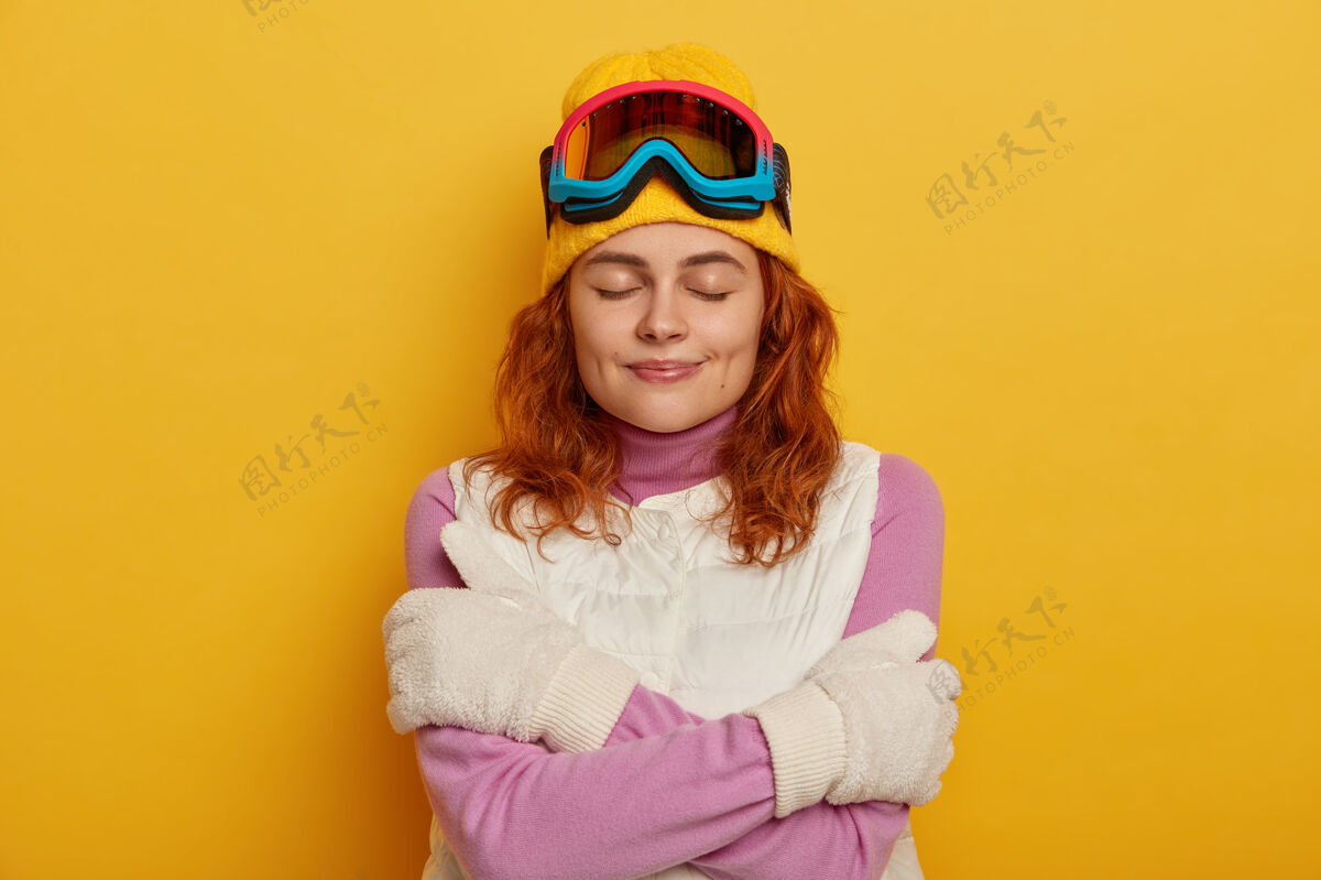 帽子漂亮的姜女在户外活动后感到寒冷 用拥抱温暖自己 戴着黄色的帽子 白色的背心和手套 闭上眼睛 隔离在黄色的背景下酒窝背心拥抱